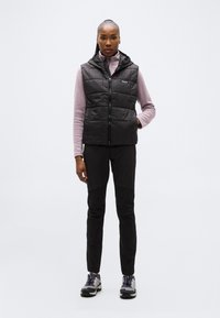 Γυναίκα όρθια, φοράει μαύρο αμάνικο μπουφάν με ραφές πάνω από ανοιχτό μωβ μακρυμάνικη μπλούζα, μαύρο παντελόνι και γκρι ορειβατικά παπούτσια, σε λευκό φόντο.