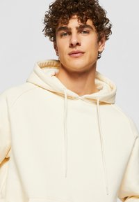 Hoodie de couleur crème en tissu doux, avec une capuche à cordon, une poche kangourou et des manches raglan.