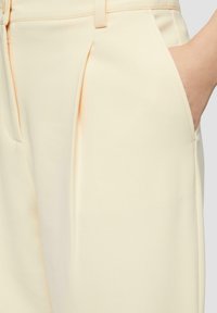 Pantalons ajustés jaune clair en tissu lisse, présentant un design à front plissé et des poches latérales pour plus de fonctionnalité.