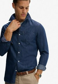 Donkerblauwe denim overhemd met een button-down kraag, borstzak en opgerolde mouwen, gecombineerd met een bruine gevlochten riem en lichtbeige broek.