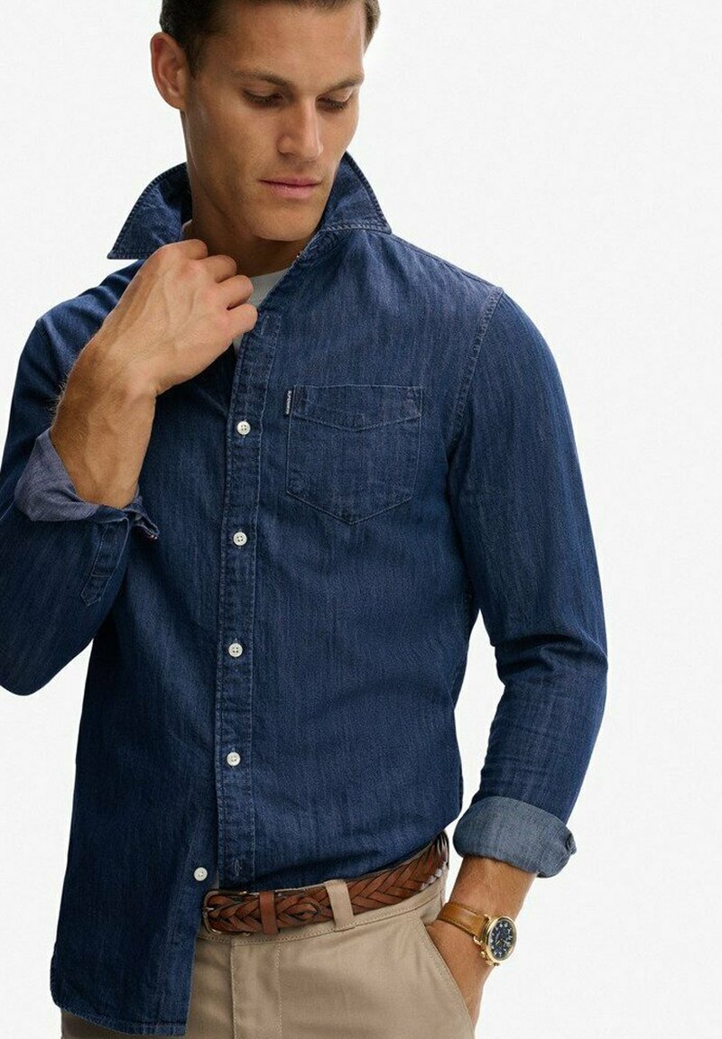 Donkerblauwe denim overhemd met een button-down kraag, borstzak en opgerolde mouwen, gecombineerd met een bruine gevlochten riem en lichtbeige broek.
