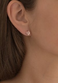 Zaželjeni uhani v obliki srčka, izdelani iz rožnatega zlata; gladka površina, poliran zaključek in subtilne konture. Nosijo jih na ušesni mečici modela.