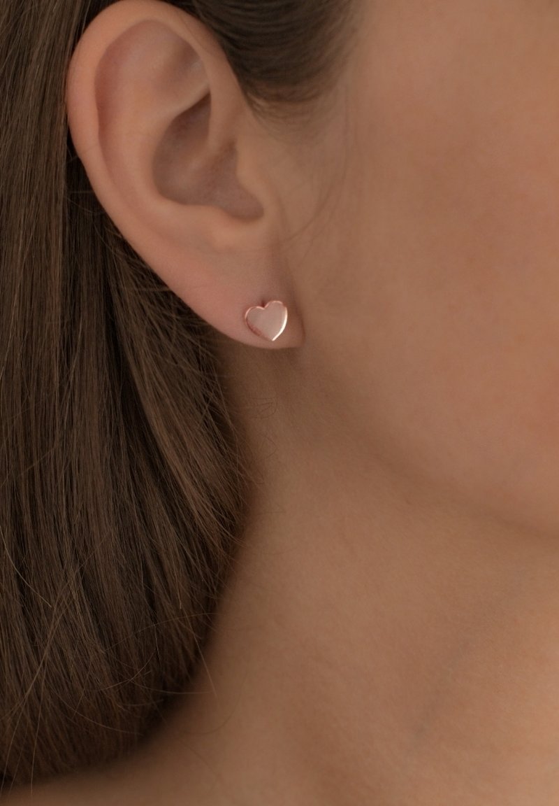 Zaželjeni uhani v obliki srčka, izdelani iz rožnatega zlata; gladka površina, poliran zaključek in subtilne konture. Nosijo jih na ušesni mečici modela.