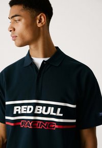Mörkblå polo-shirt i texturerad kvalitet med en vit och röd "RED BULL RACING" logotyp över bröstet, med en klassisk krage och två knappar.