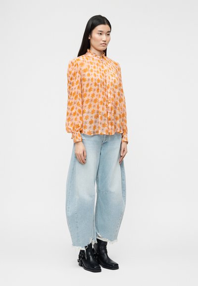 Γυναίκα στέκεται φορώντας ένα ροζ-πορτοκαλί μακρυμάνικο μπλουζάκι με λουλούδια, φαρδιά τζιν σε ανοιχτή απόχρωση με ξεφτισμένες άκρες και μαύρα μποτάκια αστραγάλου μπροστά σε μονόχρωμο φόντο.