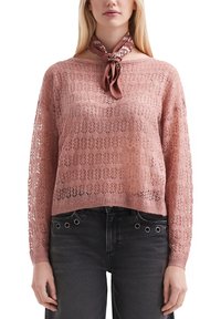 Eine Frau trägt einen staubigen rosa gestrickten Langarmpullover und schwarze Jeans mit metallenen Ösen, accessoriert mit einem gebundenen braunen gemusterten Schal.