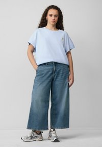 T-shirt en coton bleu clair avec un petit logo, associé à un jean en denim bleu foncé à jambe large et des chaussures de sport argentées. Design simple et décontracté.