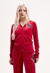 Piros velúr melegítő szett, cipzáras dzsekivel, logóval és strassz díszítéssel. Szabadidőnadrág elasztikus derékrésszel és grafikával.
