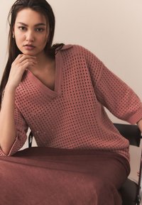 V formos iškirptės, laisvo silueto megztinis švelniai rožinės spalvos su atvira verpalų tekstūra. Dėvimas ant ilgo, rūdžių spalvos sijono. Modelis pozuoja padėjęs ranką ant smakro.