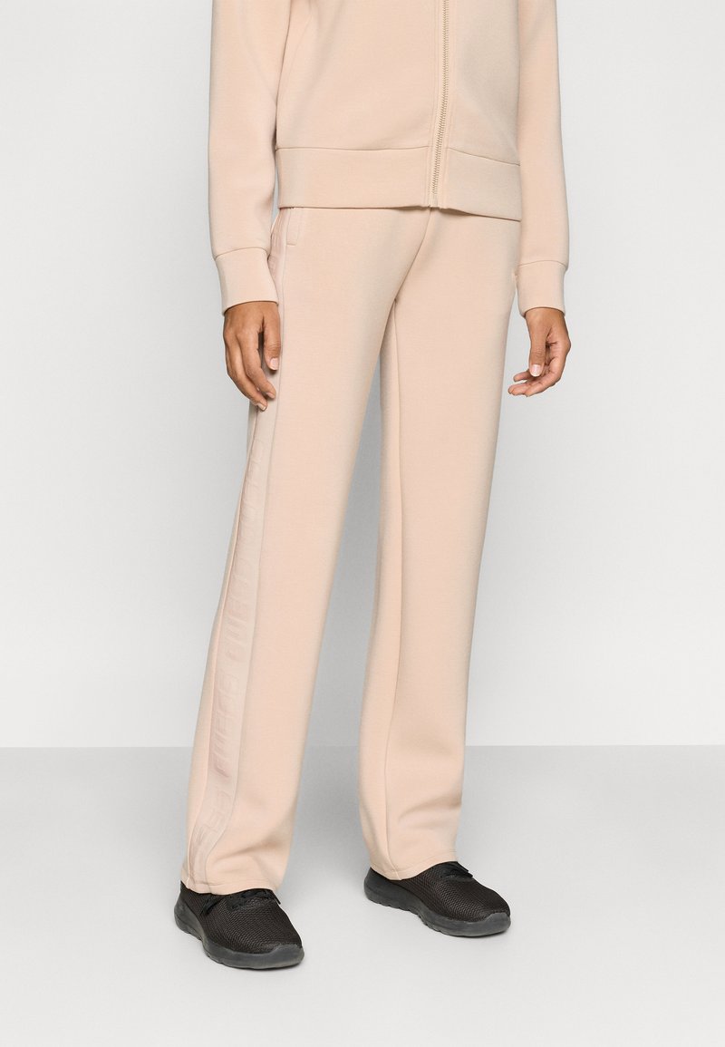 Ανοιχτό μπεζ φόρμας παντελόνι με άνετη εφαρμογή, με διακριτικό λογότυπο στο πλάι· συνδυασμένο με μαύρα αθλητικά παπούτσια. Ομαλή υφή υφάσματος.