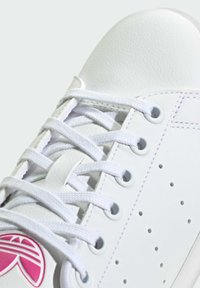 White sneakers with a textured leather upper, flat white laces, perforations for breathability, and a pink logo detail on the side.