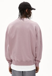 Un sweatshirt oversize rose en coton avec des poignets et un ourlet côtelés. La vue de dos montre une coupe ample et un design minimal, associé à une casquette noire.