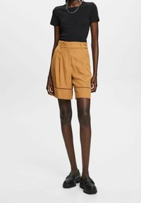 Tan hoge taille shorts met plooien en een geaccentueerde zoom, gecombineerd met een strakke zwarte t-shirt. Het model draagt zwarte instappers.