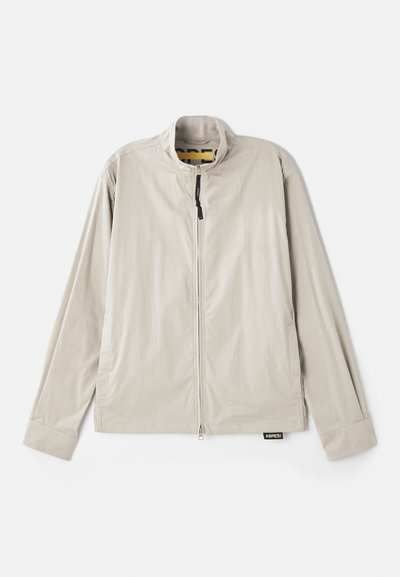 Veste beige clair avec un col haut, fermeture éclair sur le devant, et manches longues. Tissu lisse avec des poches latérales et une étiquette en bas.