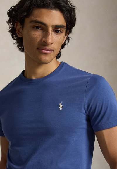 Fiatal férfi göndör sötét hajjal és szeplőkkel, kék pólót visel, amin fehér hímzett pólójátékos logó található.