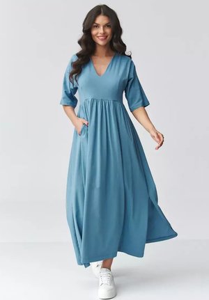 Kobieta o długich ciemnych włosach, ubrana w niebieską sukienkę maxi z dekoltem w serek i kieszeniami, w białych tenisówkach, uśmiecha się i idzie w przód na jednolitym tle.