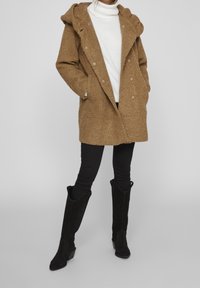 Abrigo marrón texturizado con capucha, botones de doble botonadura y bolsillos frontales, combinado con pantalones negros ajustados y botas negras hasta la rodilla.