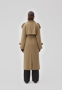 Manteau long en laine de type trench, avec une taille ceinturée, des épaulettes et des manches larges. La vue de dos montre une fente au bas.