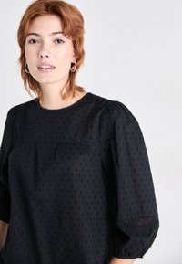 Black blouse with puffed sleeves, featuring a textured polka dot pattern and a lace accent on the yoke. Smooth fabric with a round neckline.