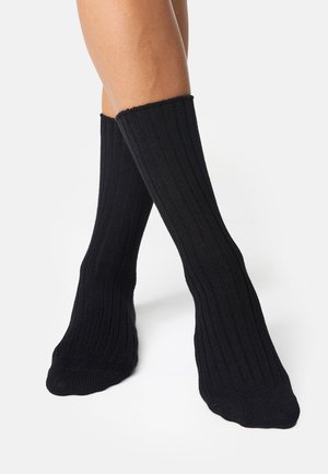 Calzini neri in maglia a coste con una texture liscia, alla caviglia, caratterizzati da un bordo spesso e una finitura morbida, indossati su una gamba parzialmente visibile.