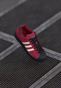 Red suede sneaker with black rubber toe cap and sole. Features white stripes and black laces. Logo on tongue and side. Textured surface.