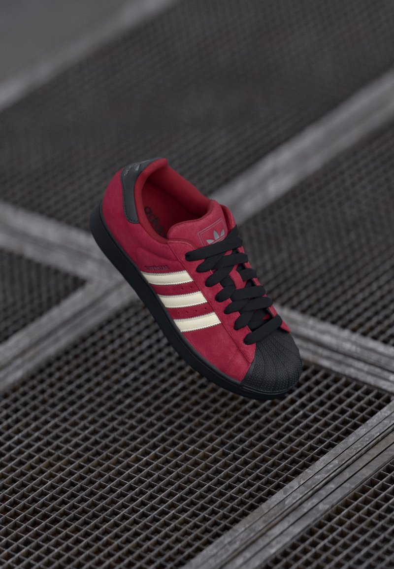 Red suede sneaker with black rubber toe cap and sole. Features white stripes and black laces. Logo on tongue and side. Textured surface.