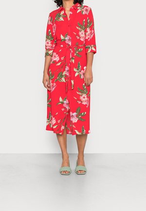 Vrouw draagt een rode midi overhemdjurk met roze bloemenprints, driekwart mouwen, een voorstrikceintuur en mintgroene gevlochten open teen hakken.