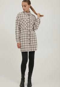 Karohemdkleid in Beige und Schwarz, mit Knopfleiste, langen Ärmeln und einem leicht lockeren Schnitt. Kombiniert mit schwarzen Skinny-Jeans und Stiefeln.