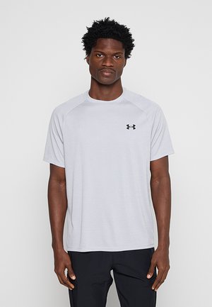 Mężczyzna stoi przodem, ubrany w jasnoszarą koszulkę Under Armour z krótkim rękawem i czarne spodnie na jednolitym tle.