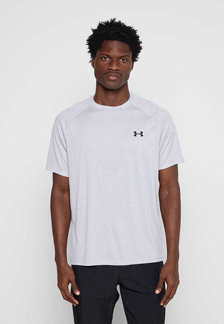 Homme debout face à l’avant, portant un t-shirt Under Armour gris clair à manches courtes et un pantalon noir sur un fond uni.