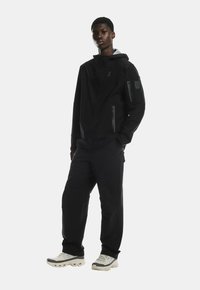 Černá bunda na zip s kapucí, se zipovými kapsami, v kombinaci s černými kalhotami a světlými sportovními botami. Hladká textura.