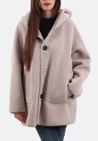 Cappotto in pile beige con cappuccio, grandi tasche frontali e due bottoni. Presenta una superficie morbida e texturizzata e una vestibilità rilassata e oversize.