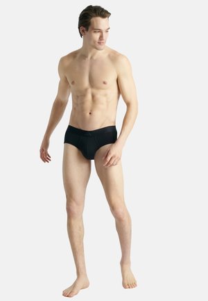 Schwarze Schwimmslips mit einfarbigem Design, elastischem Bund und passgenauer Passform, präsentiert von einem Model vor einem neutralen Hintergrund.