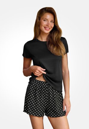 Mujer sonriendo, vistiendo una camiseta negra de manga corta y pantalones cortos negros con lunares blancos, de pie frente a un fondo liso.