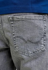 Poche arrière d'un jean en denim gris avec un motif de vague cousu, porté avec un haut en tissu texturé bleu.