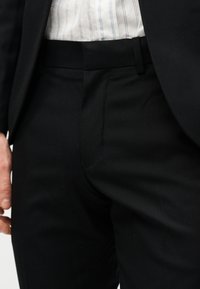 Άνδρας που φορά μαύρο παντελόνι κομμένο και ραμμένο με μαύρη ζώνη, λευκό πουκάμισο με λεπτές κάθετες ρίγες μαζεμένο μέσα και μαύρο σακάκι.