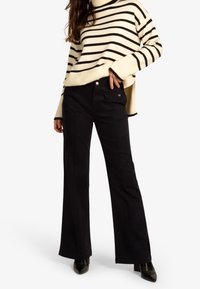 Pantalons larges noirs avec des détails de couture à l'avant et des accents de boutons, associés à un pull ivoire avec des rayures horizontales noires.