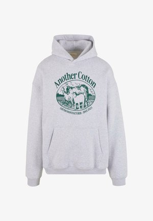 Siva bombažna kapuca z sprednjim žepom in grafičnim dizajnom, ki prikazuje kravo ter besedilo "Drugi bombaž" v temno zeleni barvi. Nastavljiva kapuca.