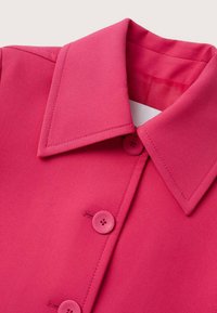 Chemise rose vif à col avec boutons assortis et couture visible sur le col et la patte de boutonnage sur un fond clair.