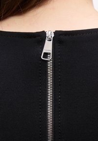 Close-up van een zilveren ritssluiting met het gegraveerde woord "Boss", vastgemaakt op de achterkant van een zwart kledingstuk gedragen door een persoon.