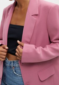 Blazer rose avec revers crantés, épaules structurées et deux boutons sur le devant. Porté sur un haut noir côtelé et un jean bleu clair.