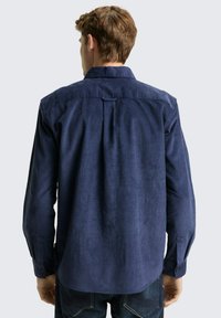 Man met kort bruin haar, gekleed in een lichtblauw corduroy overhemd met lange mouwen en donkere jeans, staande met zijn rug naar de camera tegen een effen lichte achtergrond.