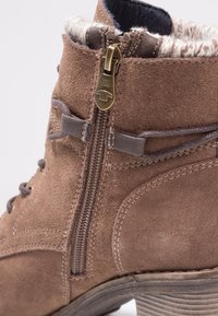 Bottine en daim marron avec fermeture éclair latérale, détails en lacets, doublure en fausse fourrure et talon texturé sur fond blanc.