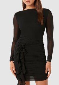 Černé šaty s dlouhým rukávem, průhlednými rukávy, nařaseným tělem, květinovým akcentem v pase a volánkovými detaily po straně.