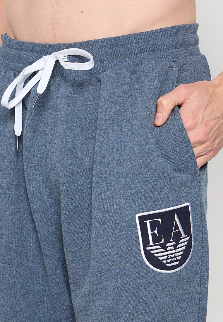 Gros plan sur une personne portant un pantalon de jogging bleu avec un cordon blanc et un logo EA marine sur la poche, la main reposant à l'intérieur de la poche.