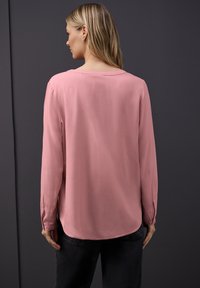 Langarmbluse aus soft-pinkem Stoff mit rundem Ausschnitt, lockerer Passform und subtiler Raffinesse, kombiniert mit schwarzen Hosen.