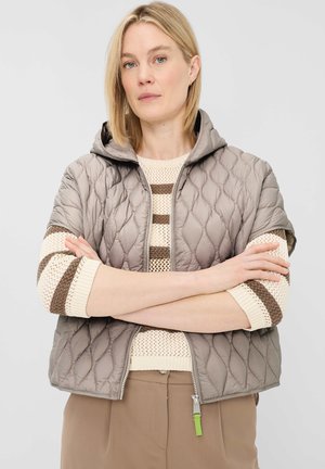 Blonde Frau mit verschränkten Armen trägt eine beige gesteppte Kurzarmjacke über einem cremefarbenen und braun gestreiften Strickpullover und beigefarbenen Hosen.