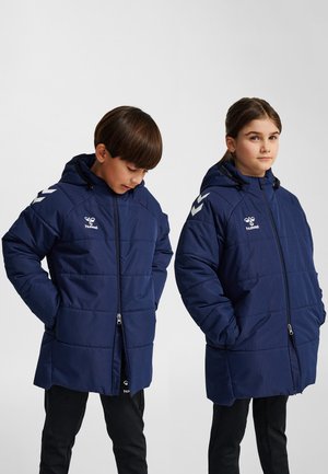 To børn iført marineblå hættetrøjer med logoer i hvid, stående med hænderne i lommerne mod en ensfarvet baggrund.