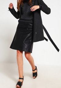 Falda negra de cuero en línea A con textura suave, combinada con un suéter negro ajustado y sandalias de plataforma negras. Abrigo largo negro sobre los hombros.