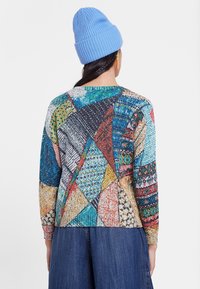 Suéter multicolorido con un diseño de patchwork que presenta formas geométricas, texturas variadas y mangas largas. Se lleva con un gorro azul.